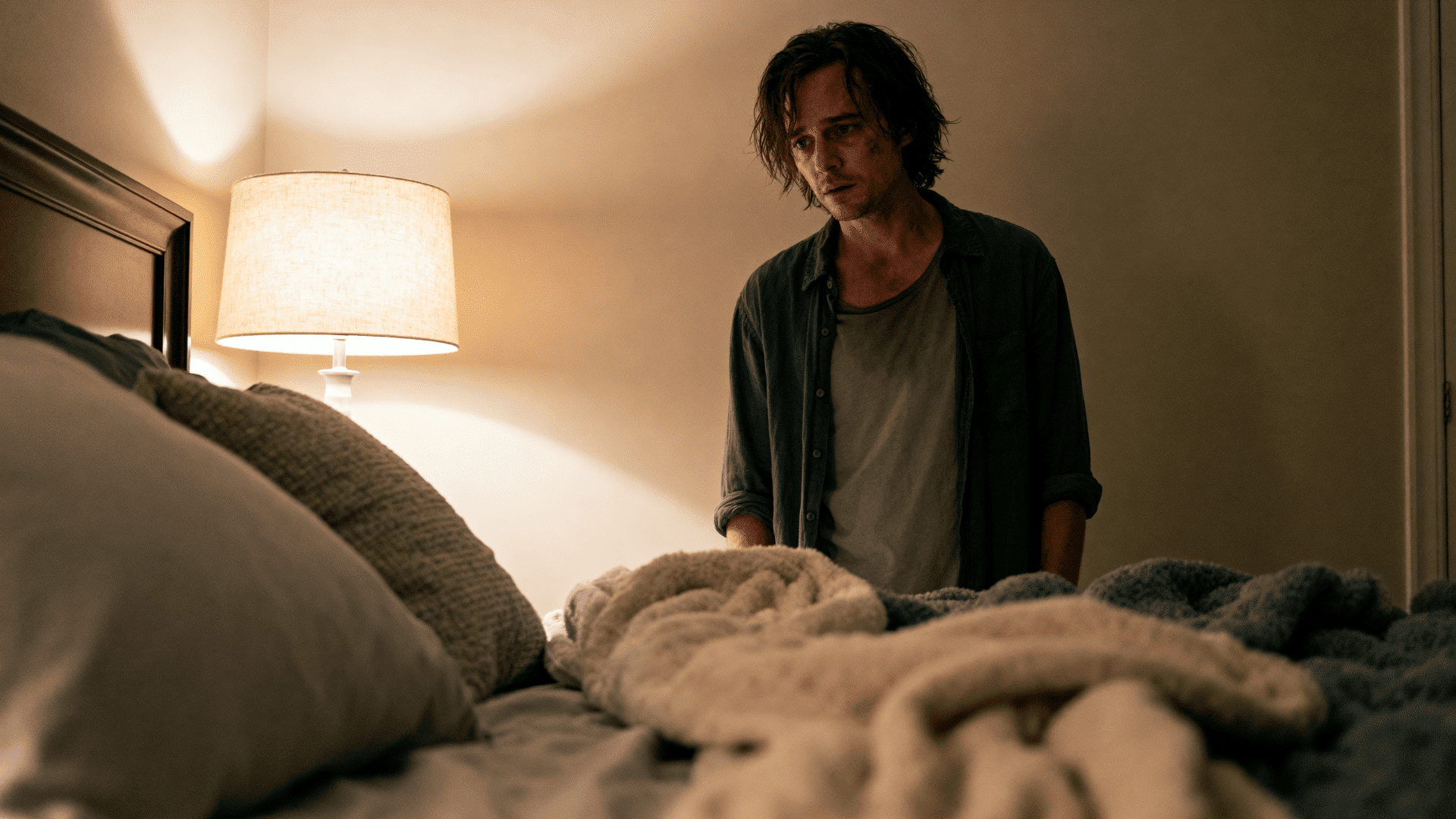Exhausted man stands beside an unmade bed in a softly lit bedroom, gazing down with tired expression under warm ambient light.