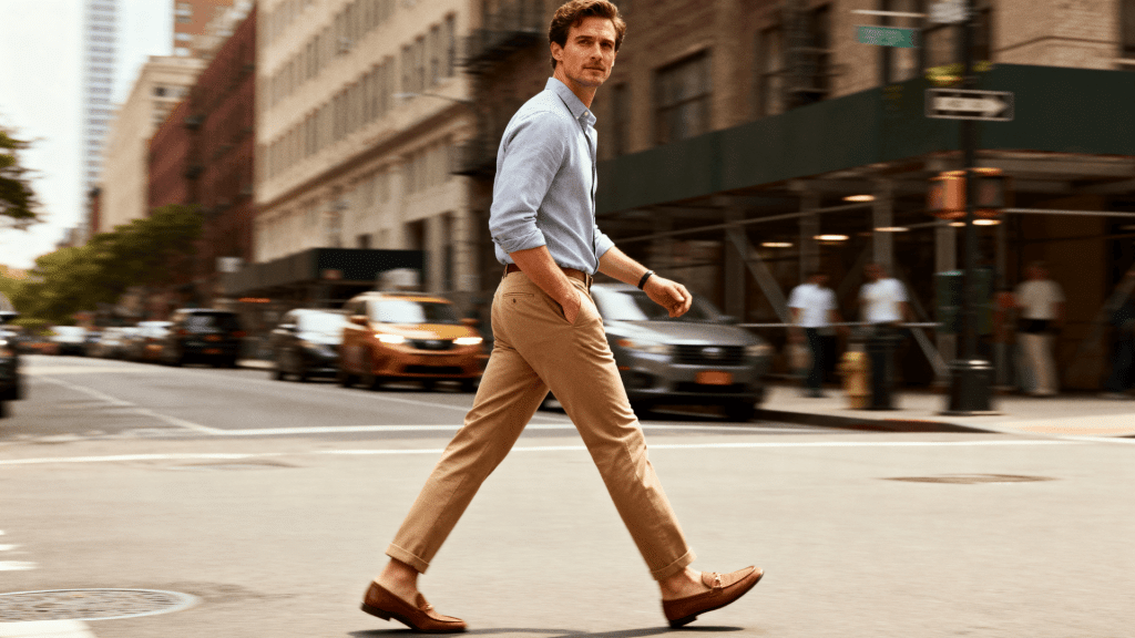 Man walking across city street wearing tan chino pants, blue shirt and loafers, styled in a smart casual urban street look