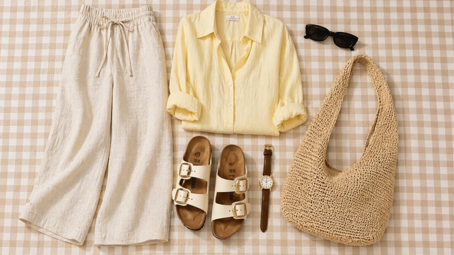 Summer flat lay with linen pants, pastel yellow shirt, sandals, sunglasses, watch, and woven bag on soft brown gingham background