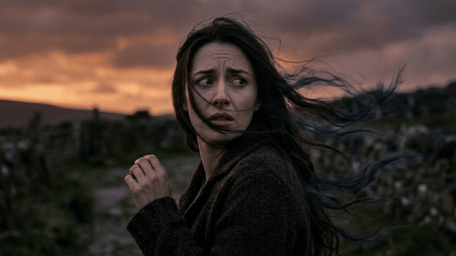 Woman with windblown hair looks back anxiously at sunset, standing in a rugged landscape with dark clouds and fading light