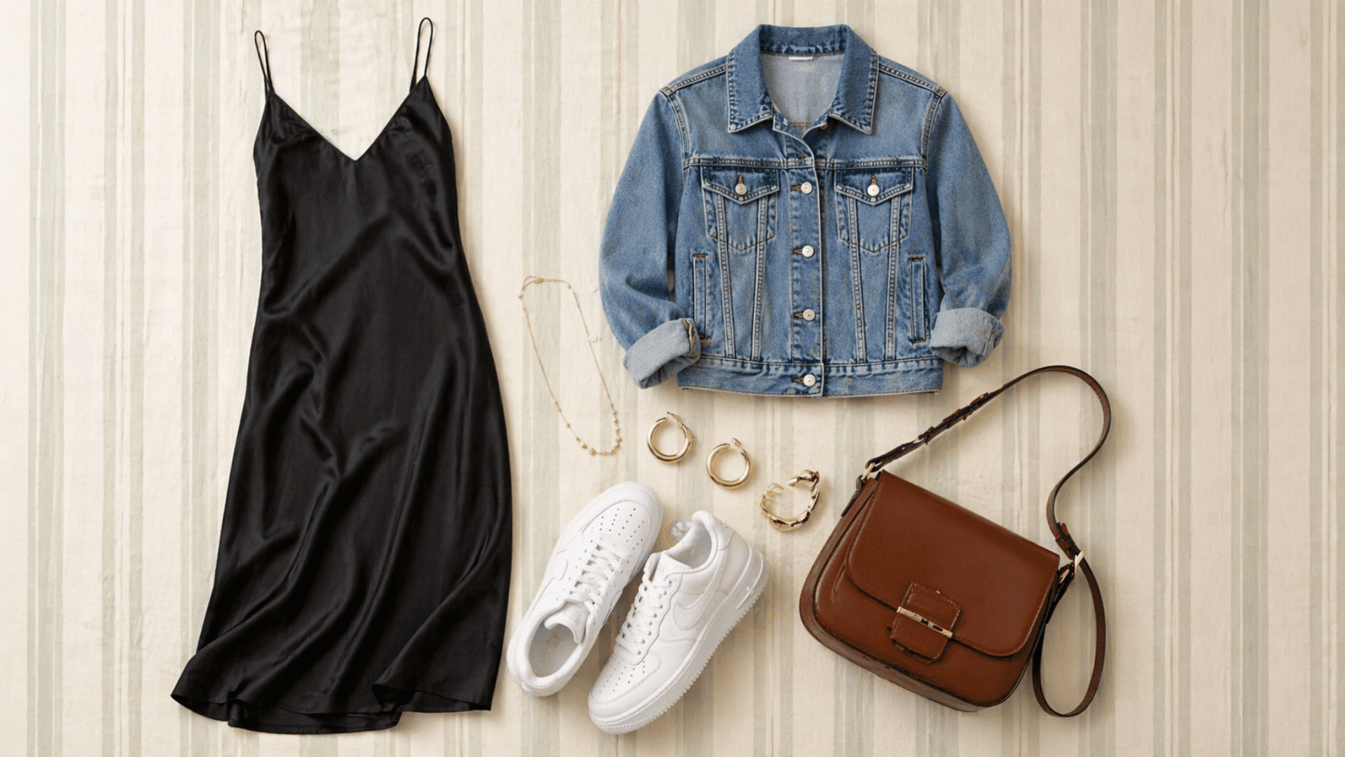 Flat lay outfit with black slip dress, denim jacket, white sneakers, brown bag, and gold jewelry on a soft striped background