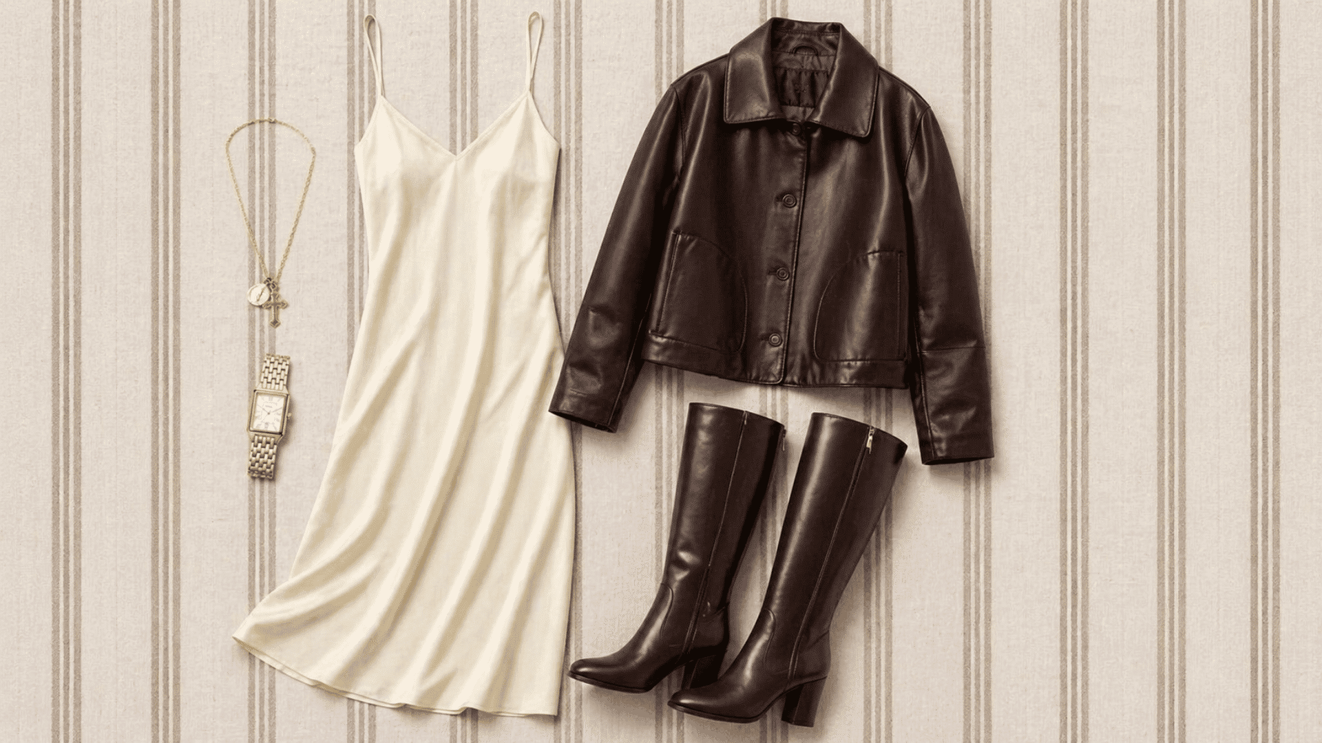 Flat lay outfit with cream slip dress, brown leather jacket, knee-high boots, gold watch and necklace on soft striped neutral background
