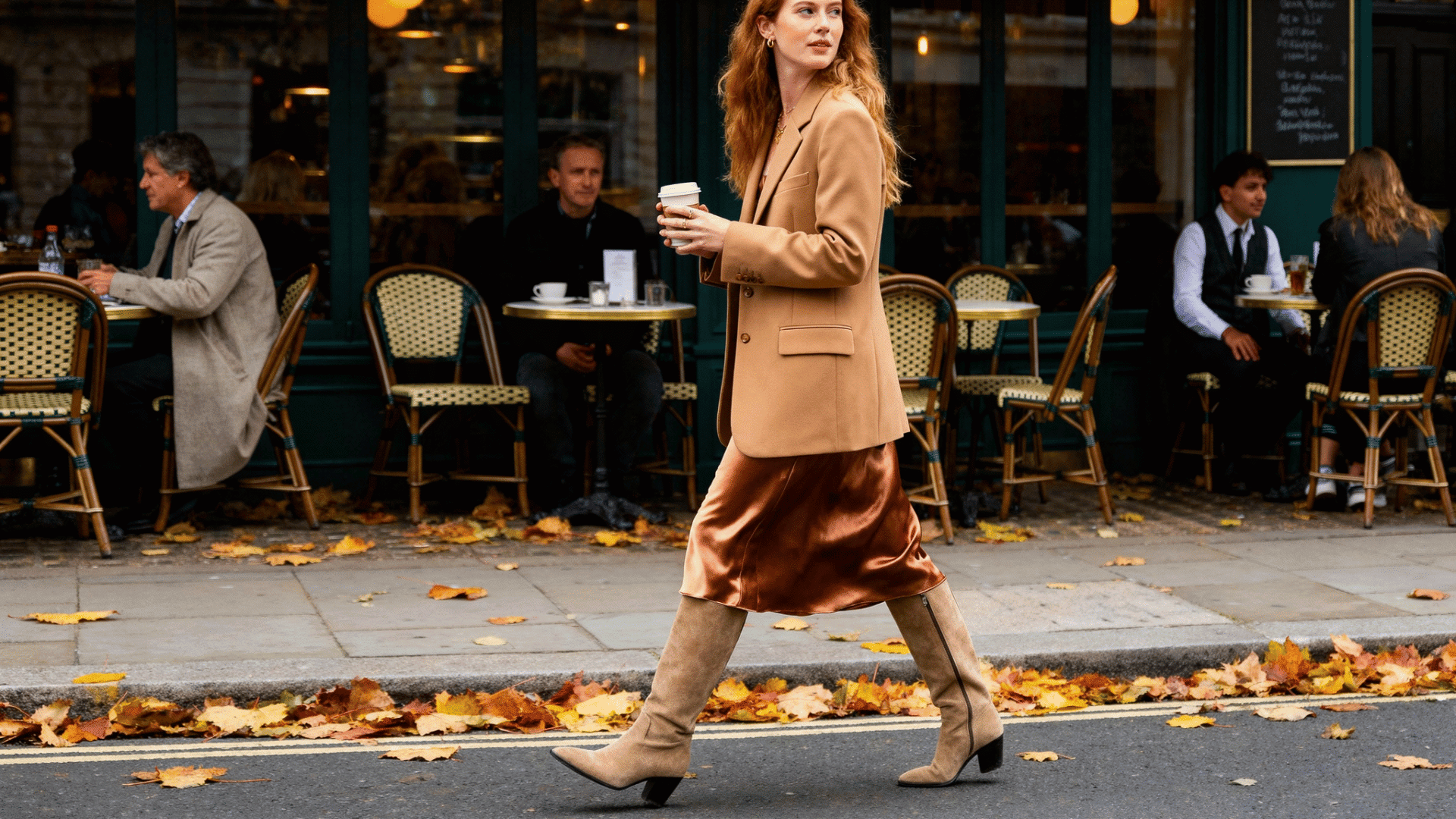 woman in a camel blazer, satin midi skirt, and knee-high boots walking past outdoor cafés on an autumn city street.