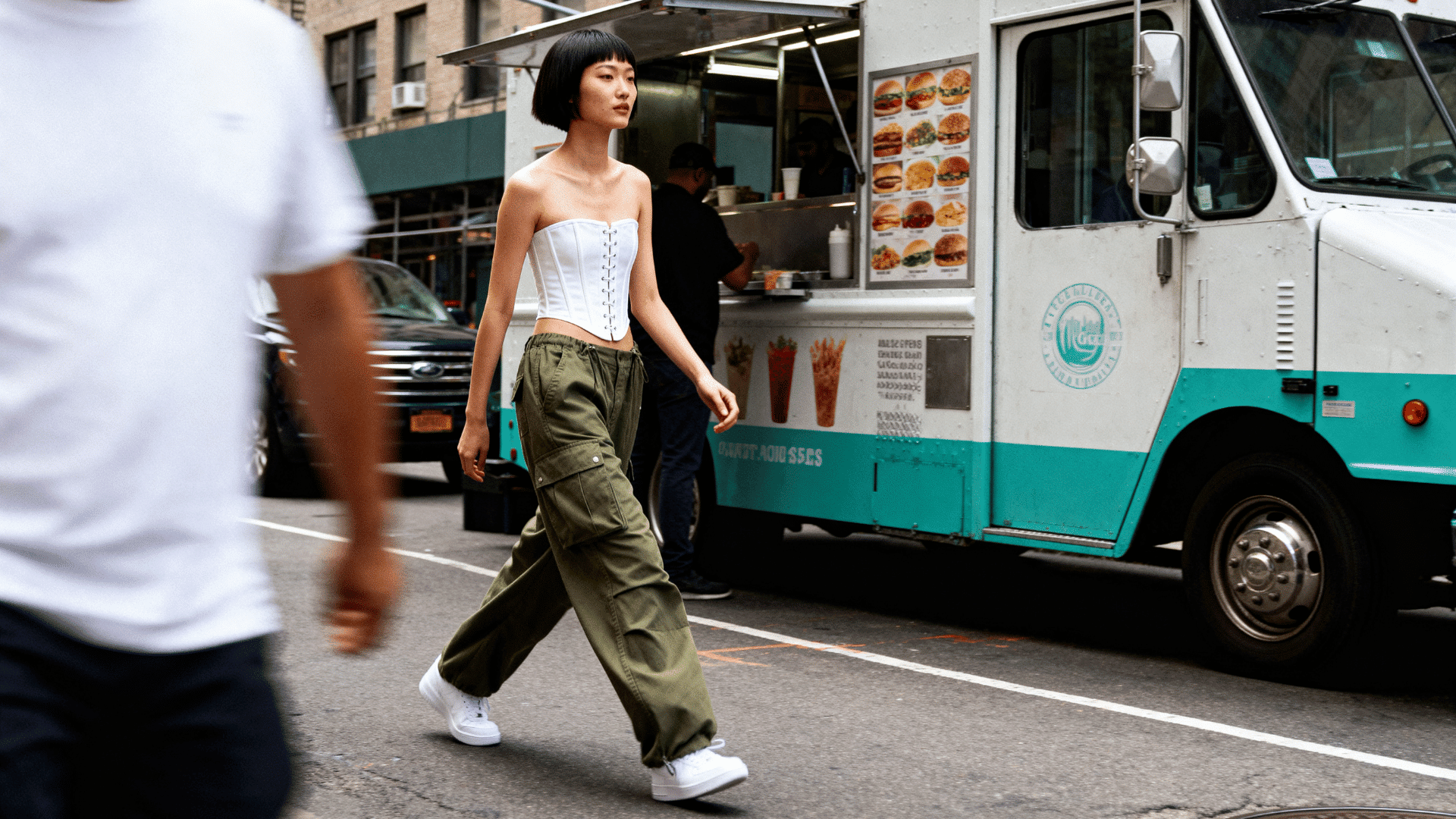 woman pairing a corset top with oversized cargo pants and sneakers on an urban street with graffiti walls and food vendors.