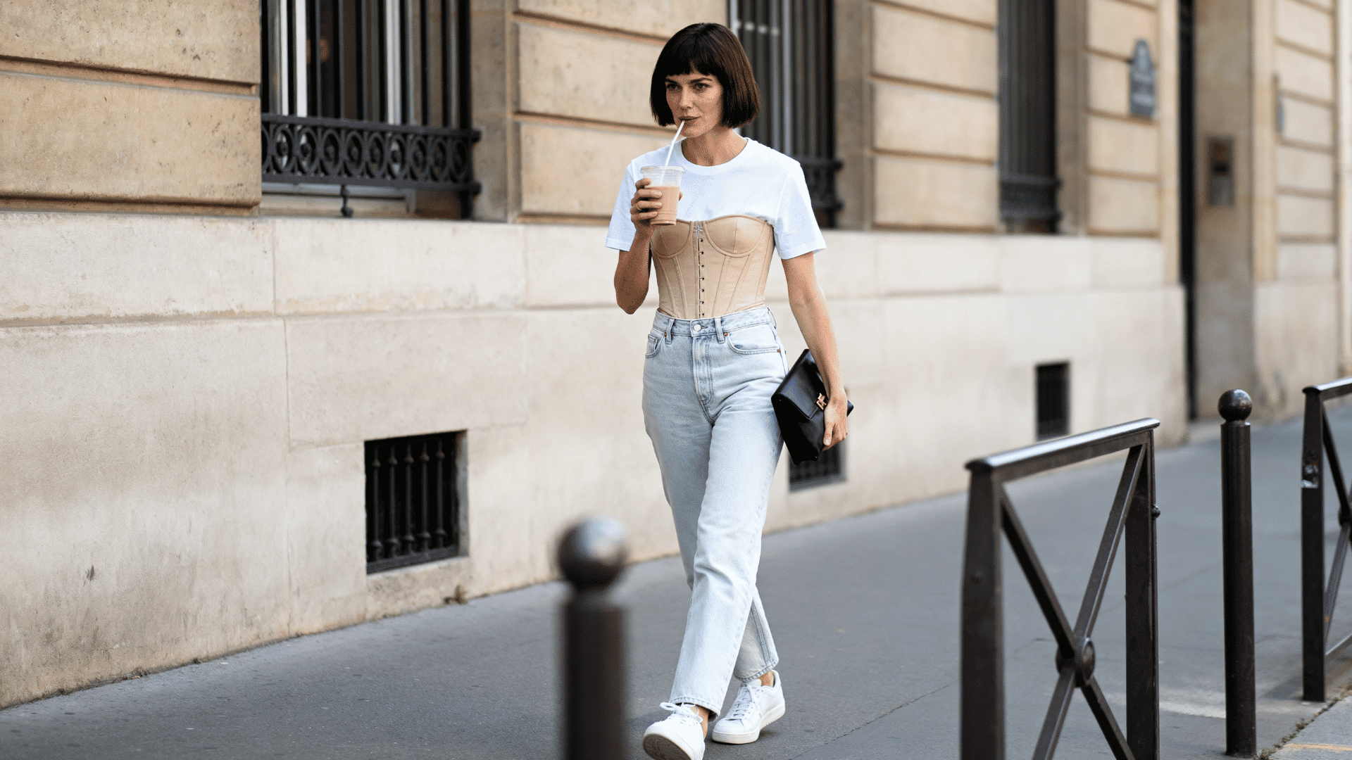 woman wearing an ivory corset top with high-waisted blue jeans and sneakers, walking along a city street with cafes and pedestrians in the background.