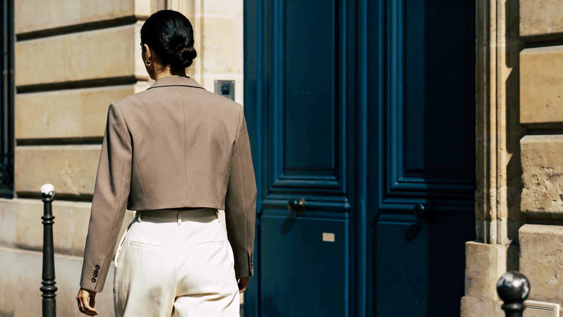 woman in a cropped blazer and cream wide-leg trousers walking outside an art gallery in a city setting.