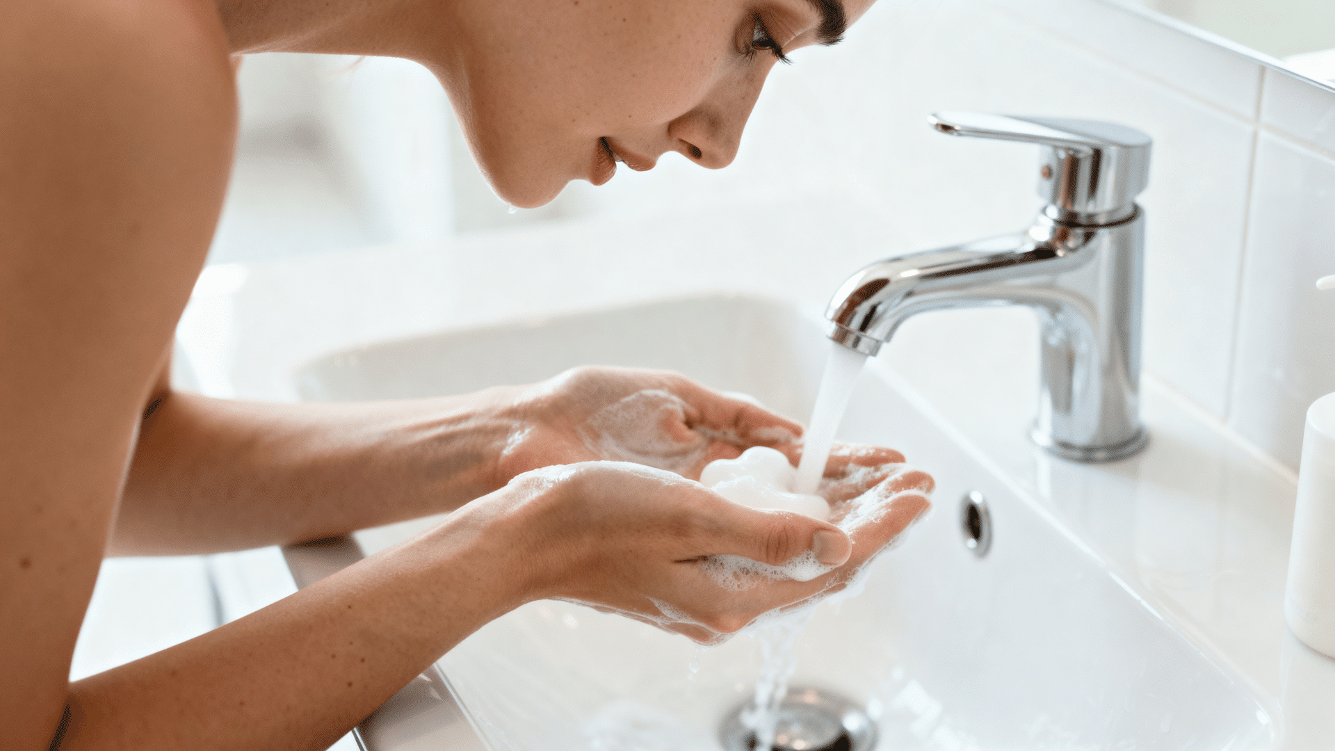 person washing their face with a gentle facial cleanser in a bright bathroom as part of a daily skincare routine