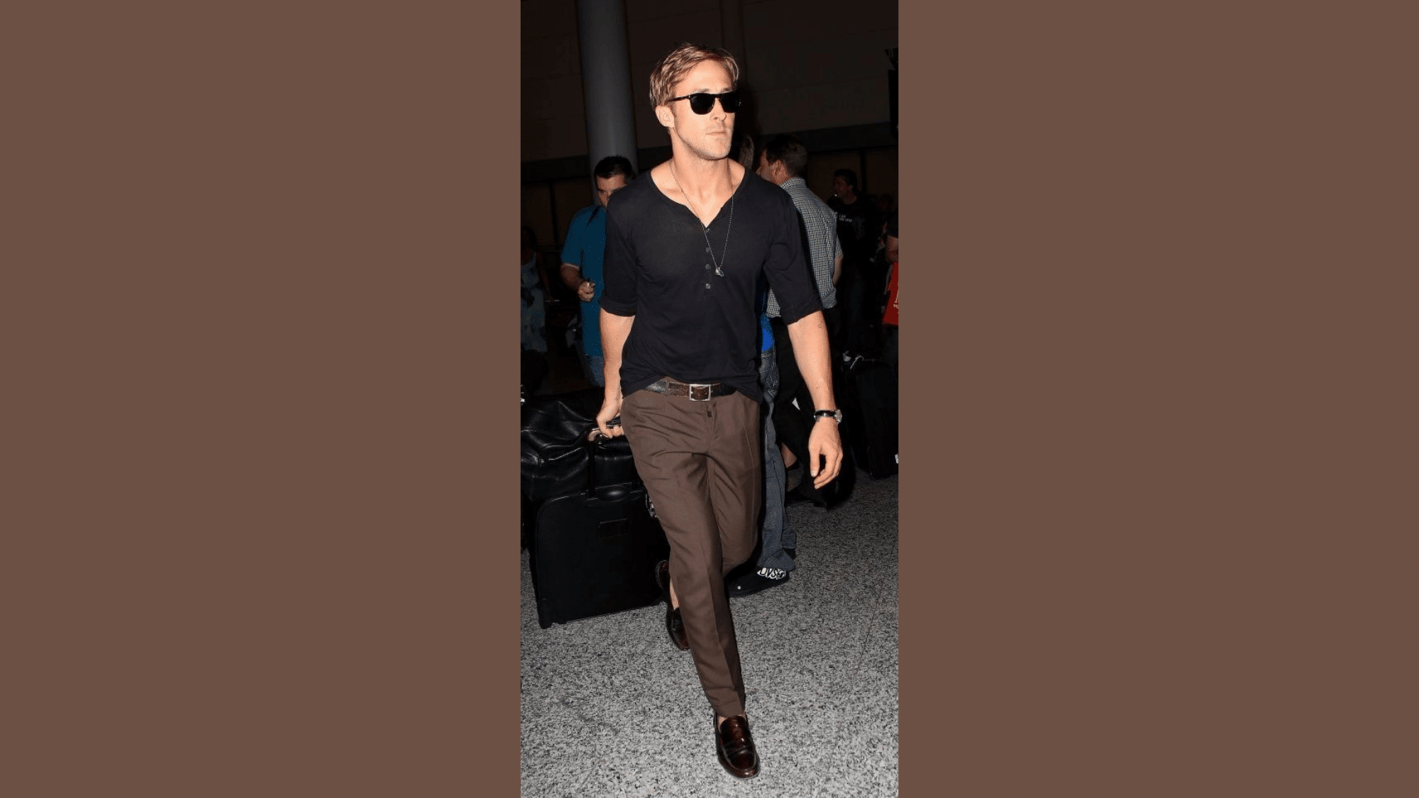 man-walking-through-airport-wearing-black-shirt-brown-pants-sunglasses-and-carrying-bag-with-people-in-background