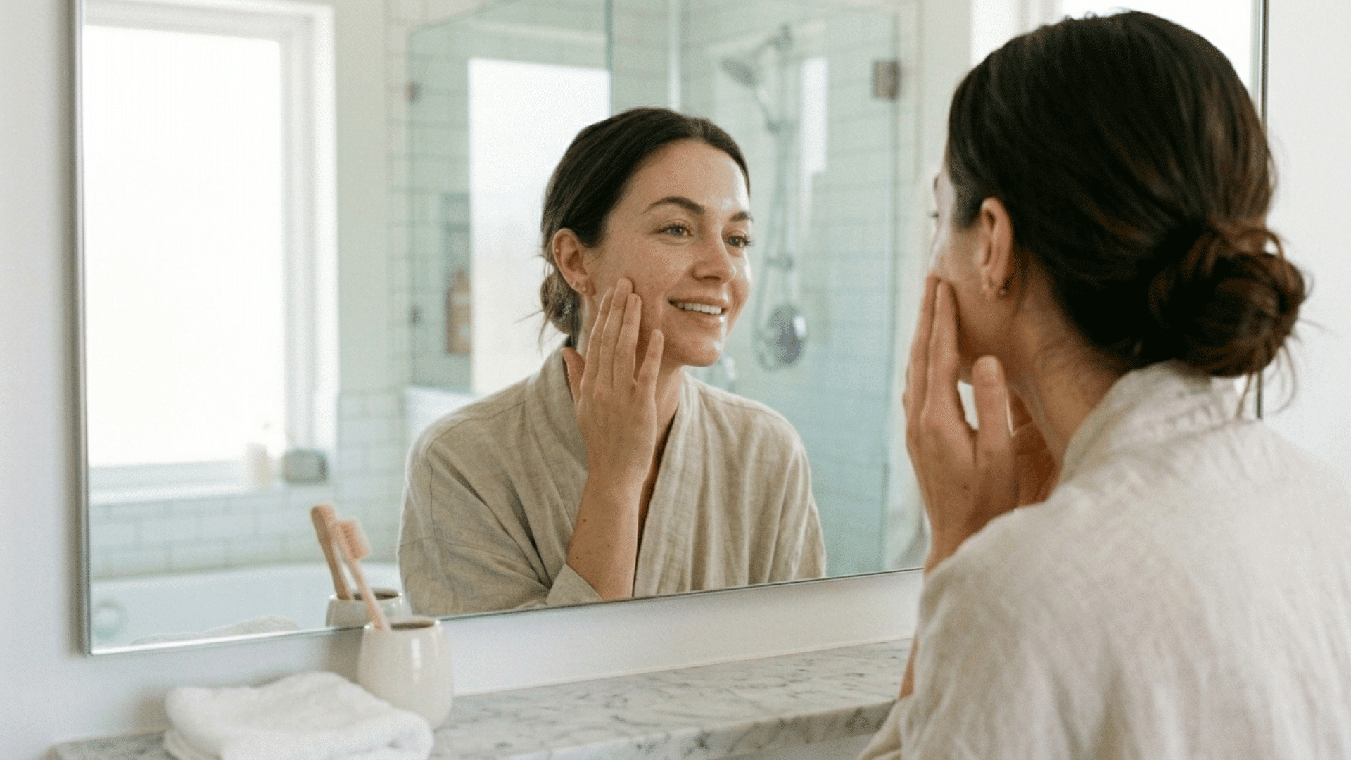 woman smiling at her reflection while gently touching her cheek in a bright bathroom during skincare routine
