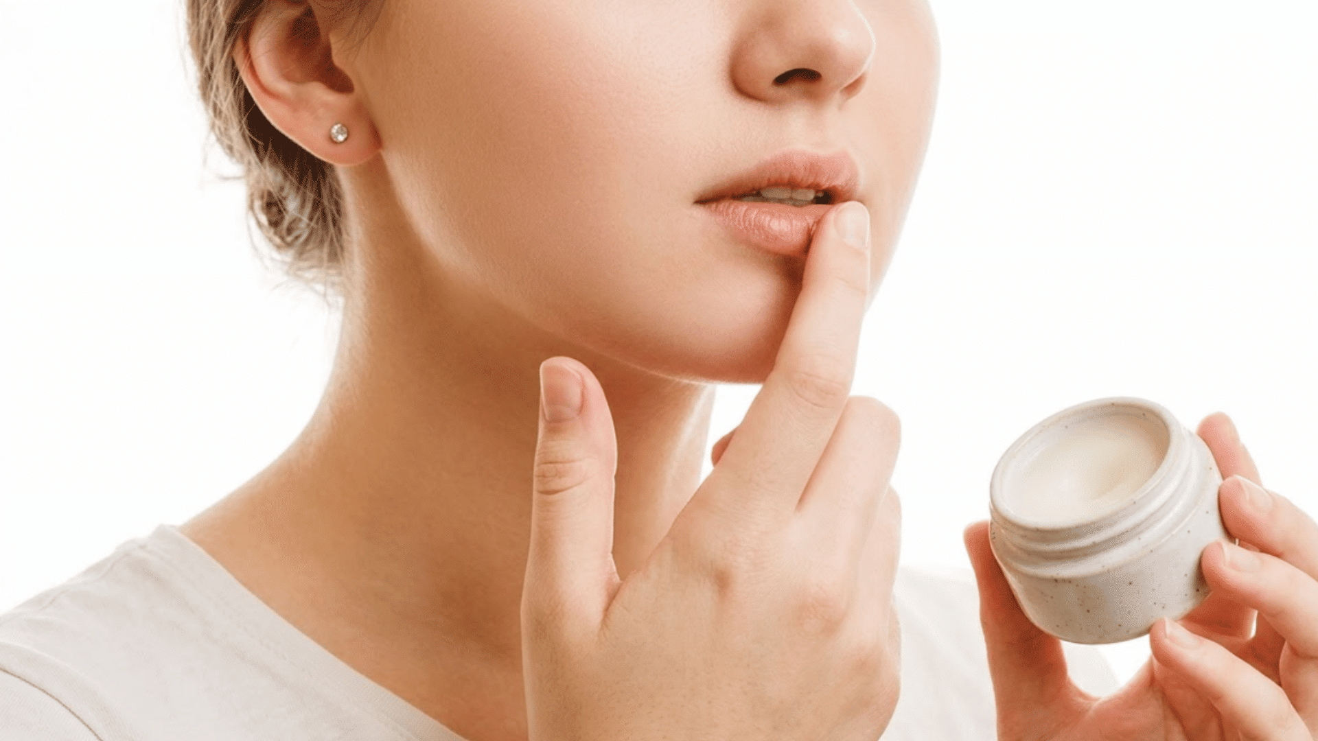 woman applying moisturizing lip balm from a small jar to dry lips against a clean white background indoors