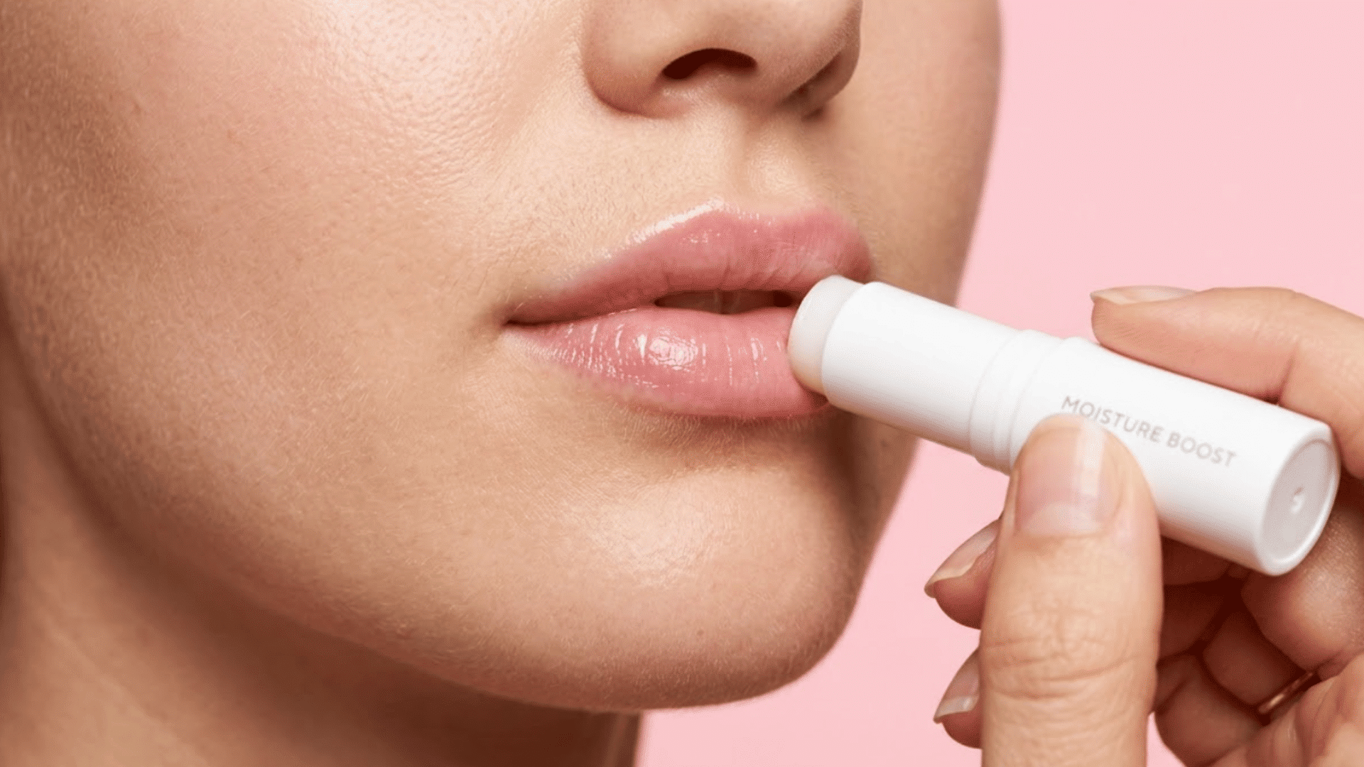 close-up of a person applying white lip balm to smooth, moisturized lips against a soft pink background