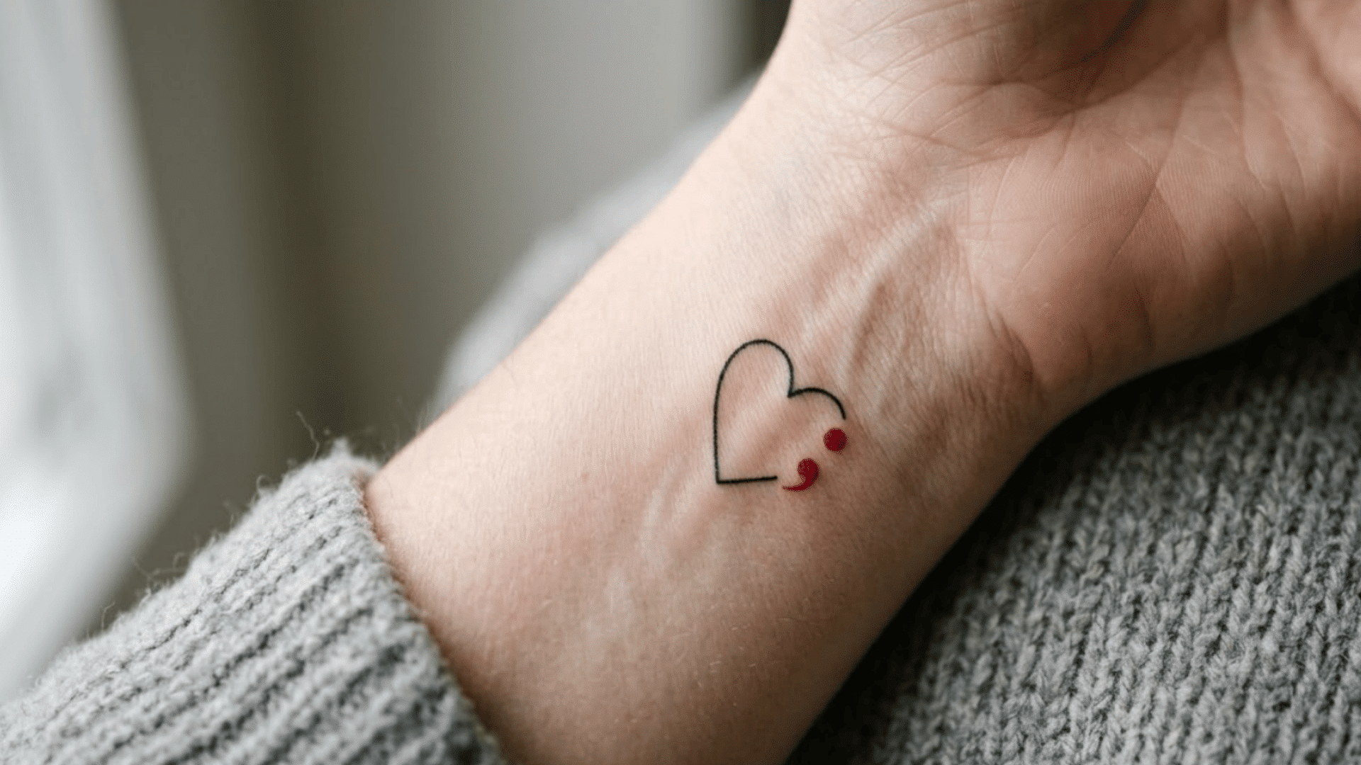 heart and red semicolon tattoo on inner wrist, shown beside a gray knit sleeve in soft natural light
