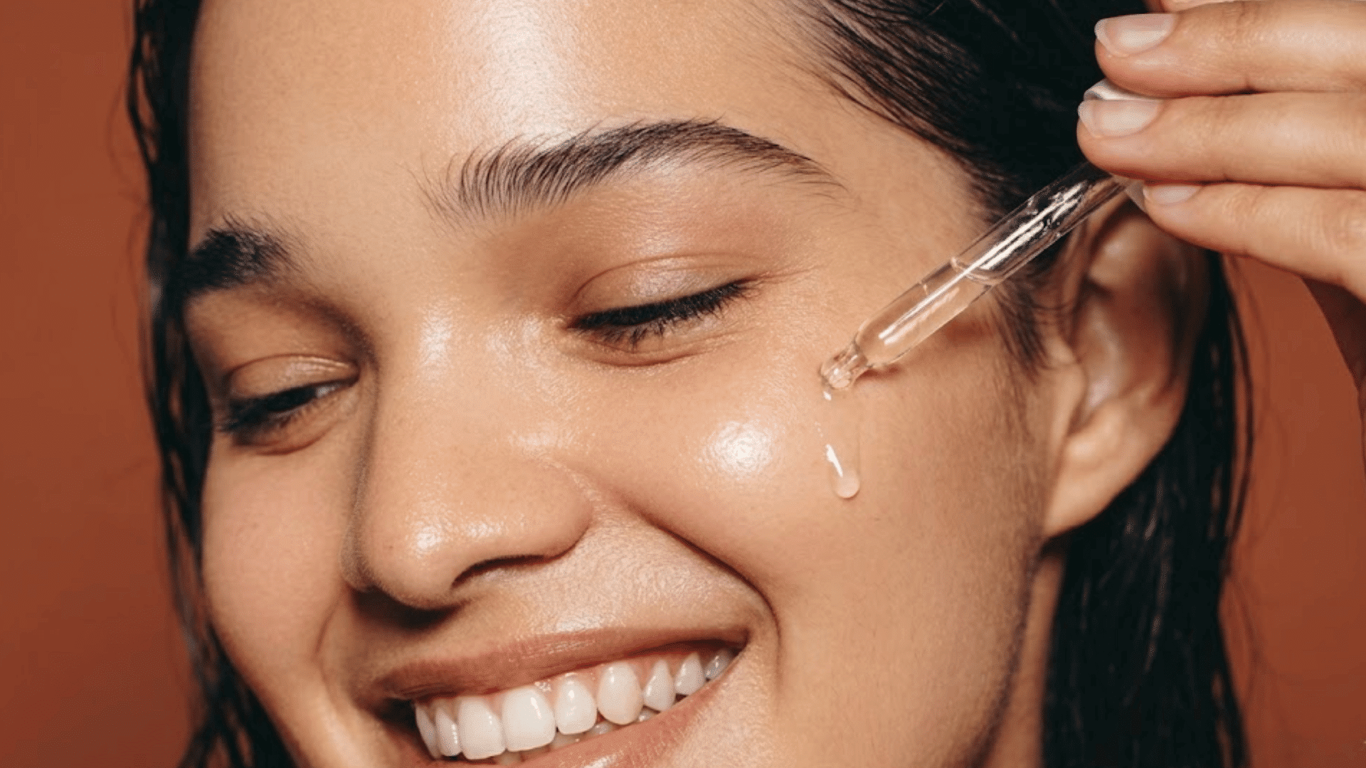 smiling woman applying hyaluronic acid serum to her cheek with a glass dropper during a skincare routine