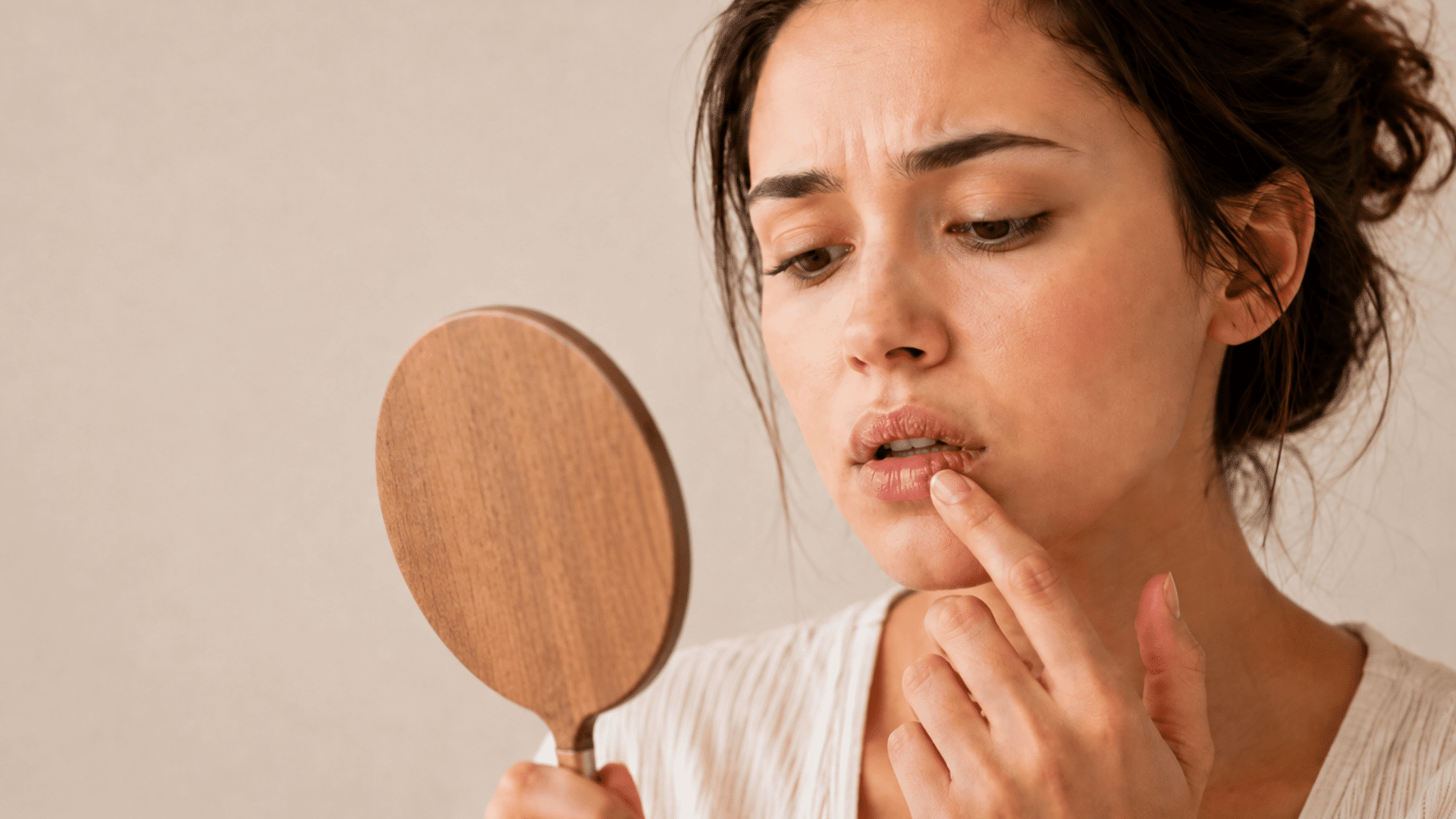 young woman checking dry peeling lips with a wooden handheld mirror against a soft beige indoor background