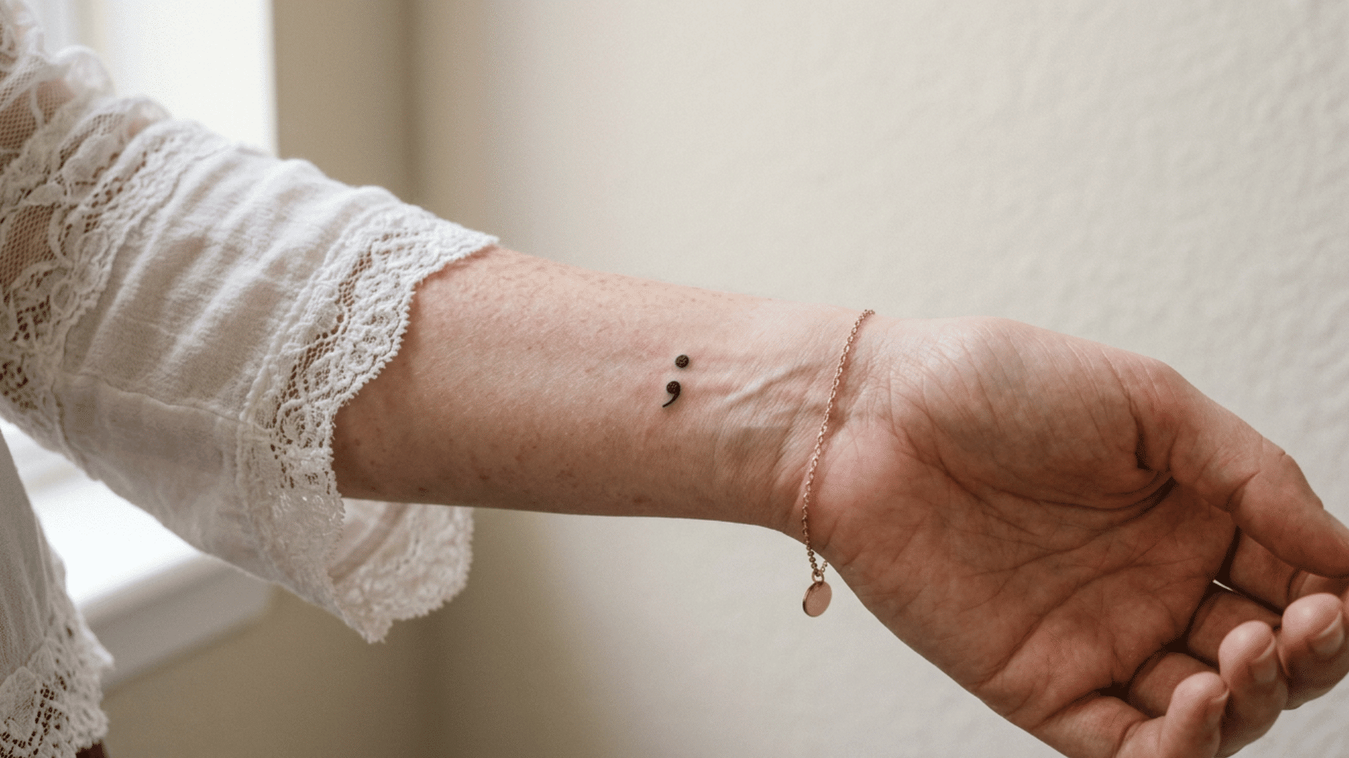 close-up of a small, minimalist black ink semicolon tattoo on the inner wrist of a person wearing a lace top