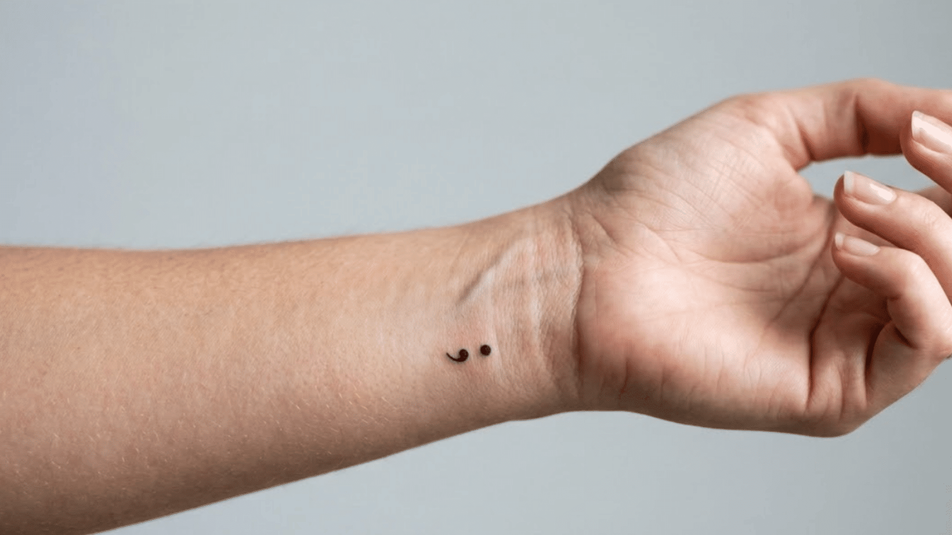 small black semicolon tattoo on inner wrist, shown against a plain gray background with a relaxed hand