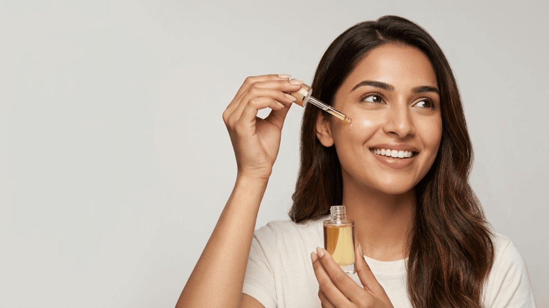 woman applying facial serum with a dropper while holding the bottle against a light neutral background