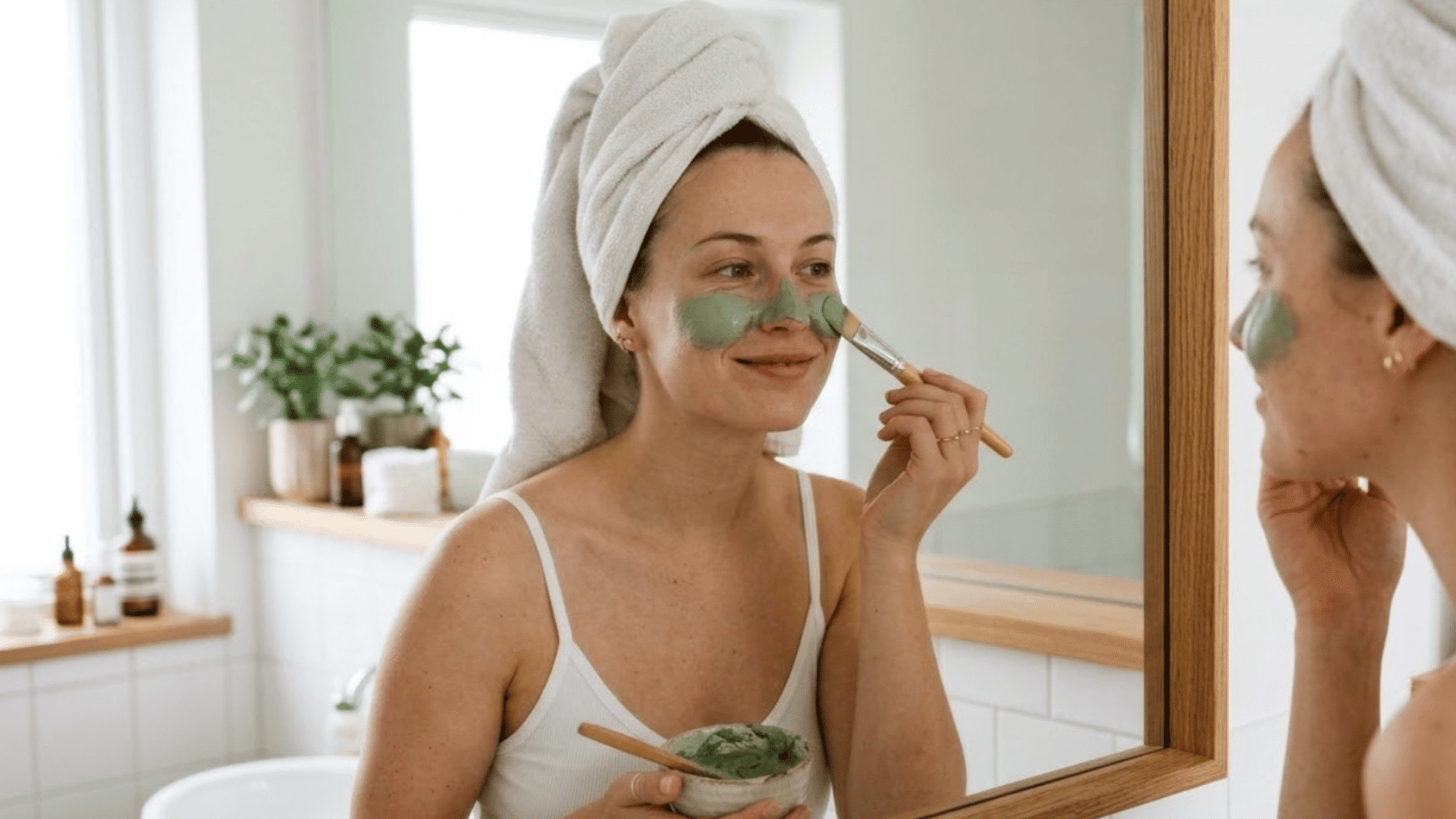 woman applying a green clay face mask in front of a bathroom mirror as part of a self-care routine